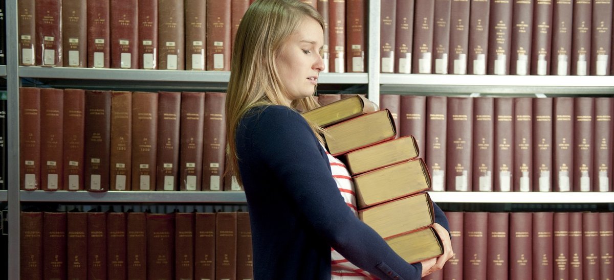 Studierende mit einem Stapel Bücher vor einem Regal voll mit Büchern.