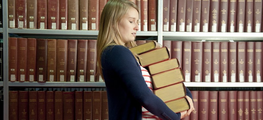 Studierende mit einem Stapel Bücher vor einem Regal voll mit Büchern.