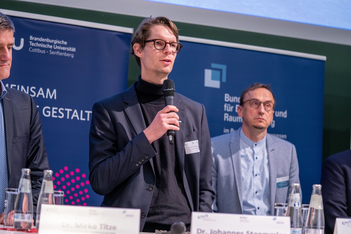 Drei Personen auf einer Bühne hinter einem Pult in einem Setting für Diskussionsrunden. Dr. Johannes Staemmler spricht am Mikrofon.