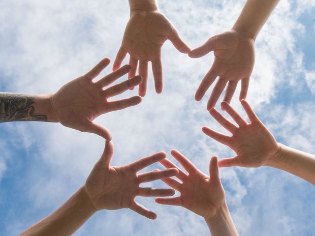 The photo shows 6 hands pictured in front of the sky, forming a circle.