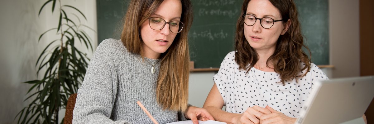 2 Frauen sitzen an einem Tisch. Eine schreibt etwas auf, während die andere spricht.