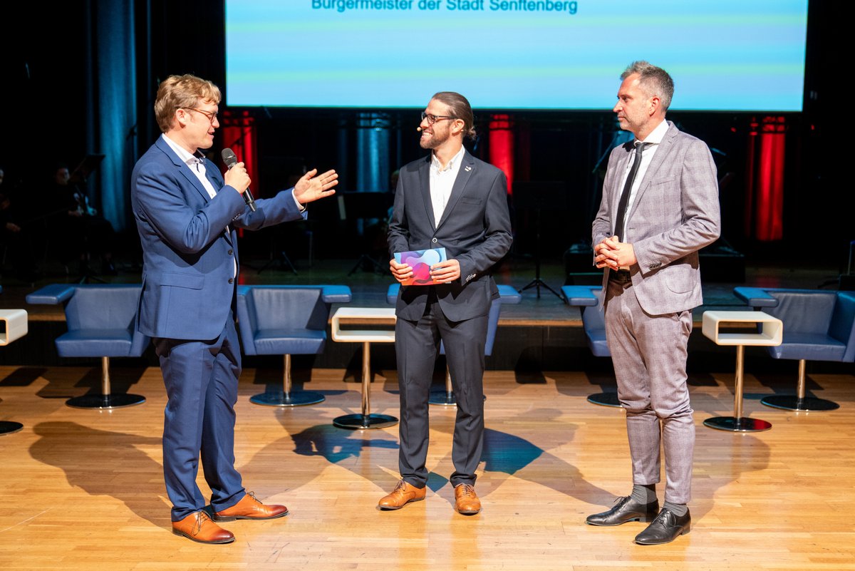 [Translate to Englisch:] Moderator Alexander Telle mit den Bürgermeistern der beiden Stadte auf der Bühne