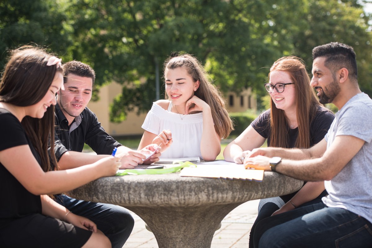 Studierende diskutieren, an einem Betontisch auf dem Campus sitzend, miteinander.