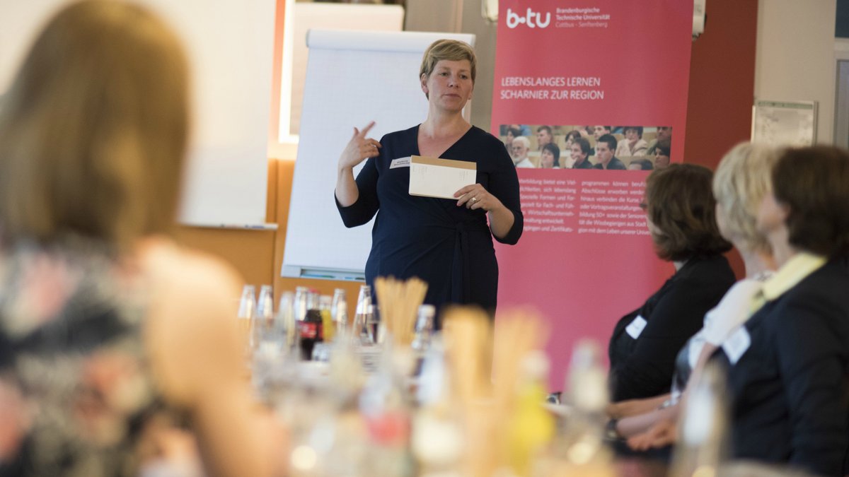 Eine Dozentin hält ein Seminar zum Thema "Lebenslanges lernen - Scharnier zur Region" mit zahlreichen Teilnehmenden ab.