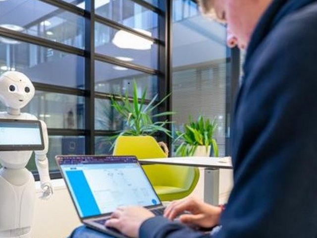 Male person working on a laptop, in front of a humanoid robot.