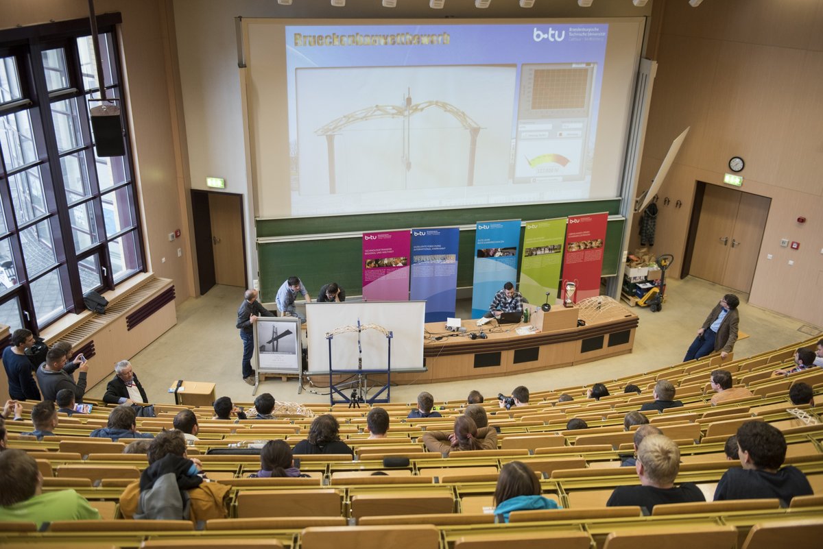 Blick in den großen Hörsaal des Konrad-Zuse-Medienzentrums während des traditionellen Brückenbauwettbewerbs. Diie Mehrzahl der Beteiligten und die Zuschauer siten in den Reihen und verfolgen, wie die Belastbarkeit einer Brücke auf dem Prüfstand getestet wird.