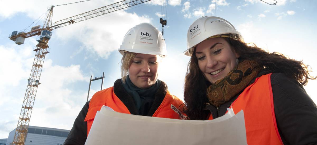 Zwei Masterstudentinnen im Studiengang Bauingenieurwesen stehen mit Bauhelmen auf der Baustelle und betrachten eienn Bauplan