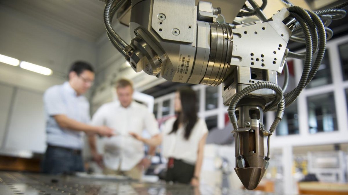 Detailansicht eines Roboters, im Hintergrund stehen drei Personen, die sich unterhalten