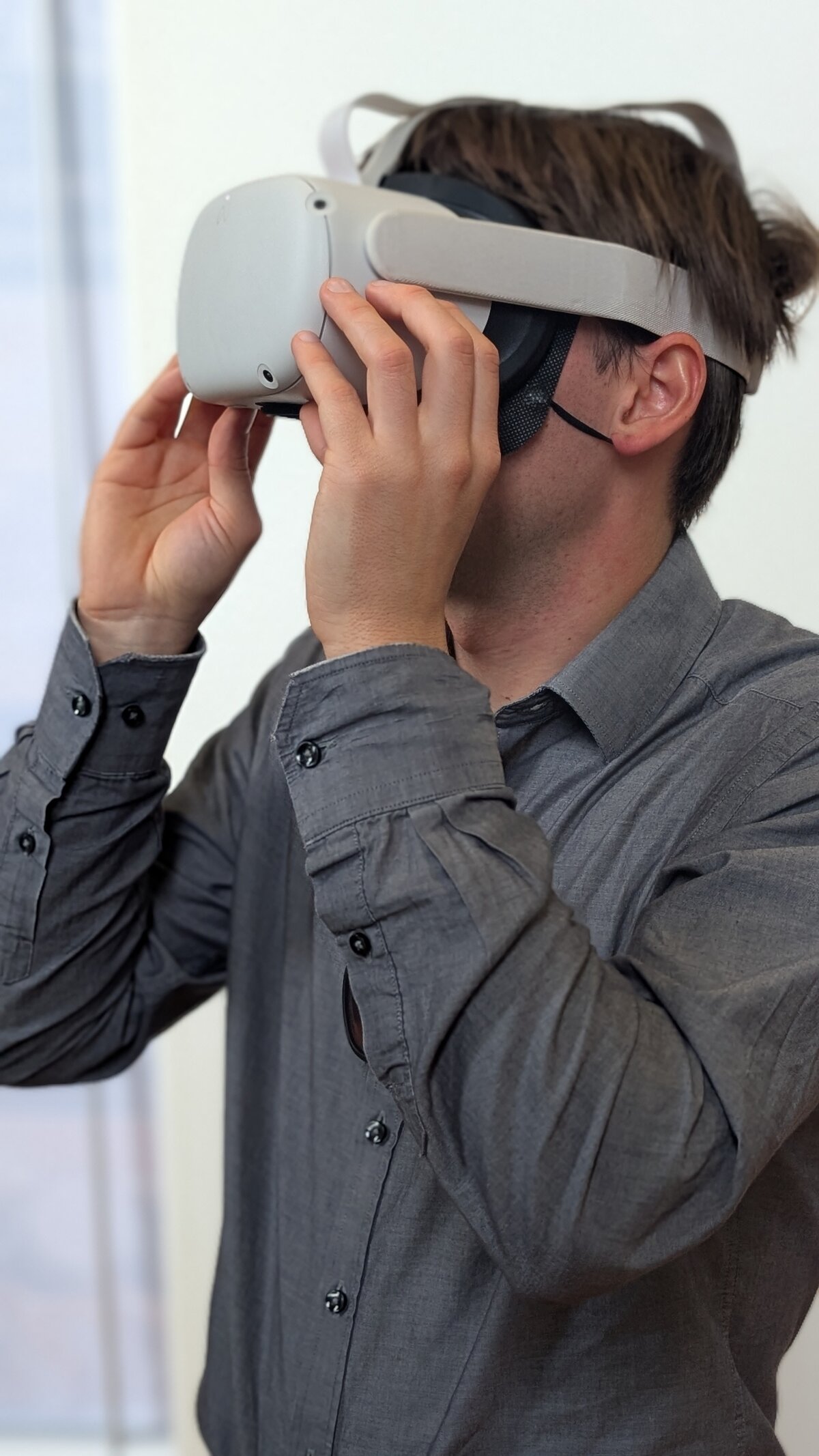 Ein Junger Mann mit grauen hemd hat eine VR-Brille auf un blickt nach links. Die Hände leicht an der VR-Brille.