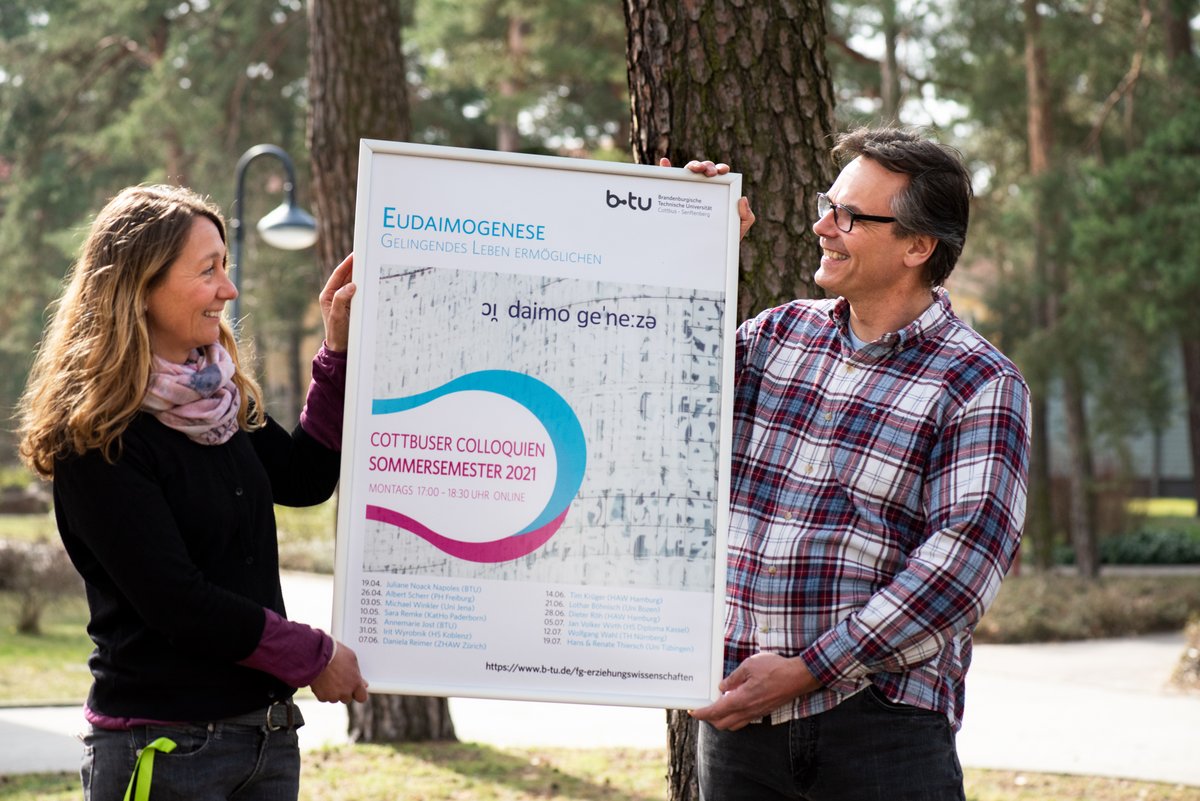 Juliane Noack Napoles und Thorsten Heimann halten gemeinsam das Plakat zur Veranstaltung "Eudaimogenese - Gelingendes Leben ermöglichen"