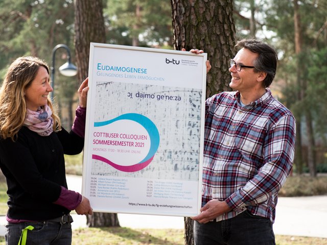 Juliane Noack Napoles und Thorsten Heimann halten gemeinsam das Plakat zur Veranstaltung "Eudaimogenese - Gelingendes Leben ermöglichen"