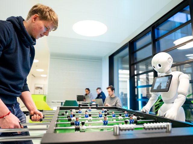 Student am Kickertisch mit einem Roboter, im Hintergrund weitere Studierende an Laptops