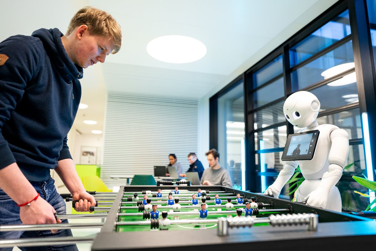 Student am Kickertisch mit einem Roboter, im Hintergrund weitere Studierende an Laptops