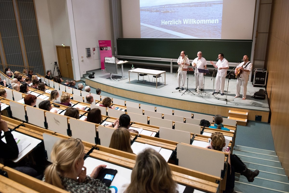 Blick in den Hörsaal mit den Teilnehmenden, auf dessen Bühne das Quartett "Diie pflegeleichten Vier" musikalisch auf die Tagung einstimmt.