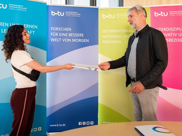 BTU-Vizepräsident Prof. Dr. Matthias Koziol übergibt, vor BTU-Bannern im Senatssaal stehend, das ESiSt-Zeugnis an die Absolventin des Projektes Roaa Mustafa.
