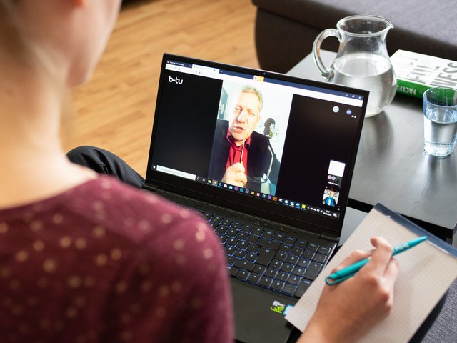 Eine weibliche Person, mit dem Rücken zum Betrachter, arbeitet im Rahmen einer Videokonferenz an einem Laptop. 
