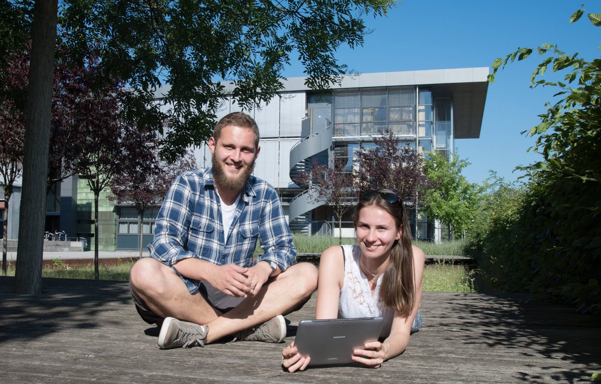 Eine Studentin und ein Student chatten mit einem Laptop auf dem grünen BTU-Campus. 