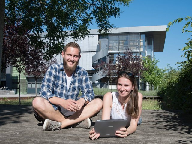 Eine Studentin und ein Student chatten mit einem Laptop auf dem grünen BTU-Campus. 