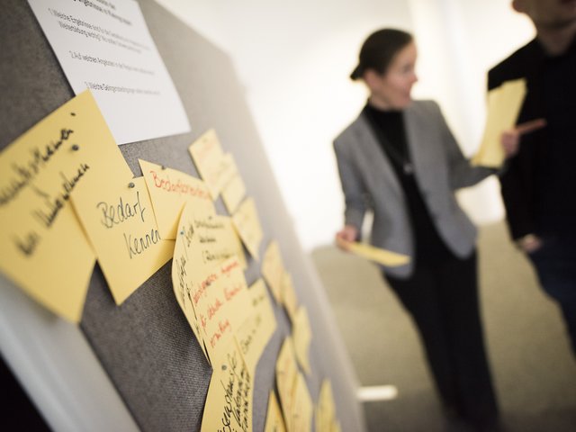 Pinwand mit Ideen, im Hintergrund Personen, die diskutieren 