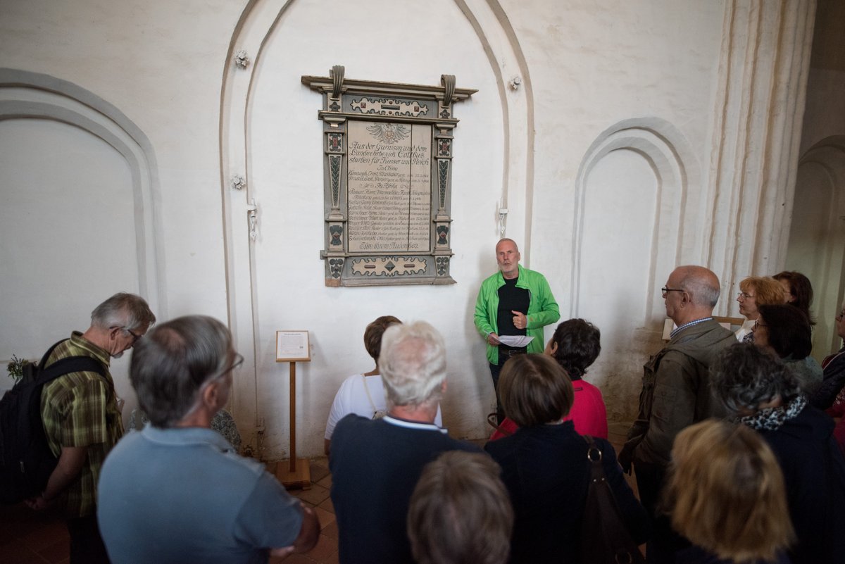 Auf dem Bild ist eine Gruppe Menschen zu sehen, die vor einer weißen Wand mit einer Gedenktafel stehen. Auf der Gedenktafel steht: "Aus der Garnison und dem Landwehrbezirk Cottbus starben Kaiser und Reich In China". Ein Mann mit Zetteln in der Hand spricht zu den restlichen Gruppenmitgliedern