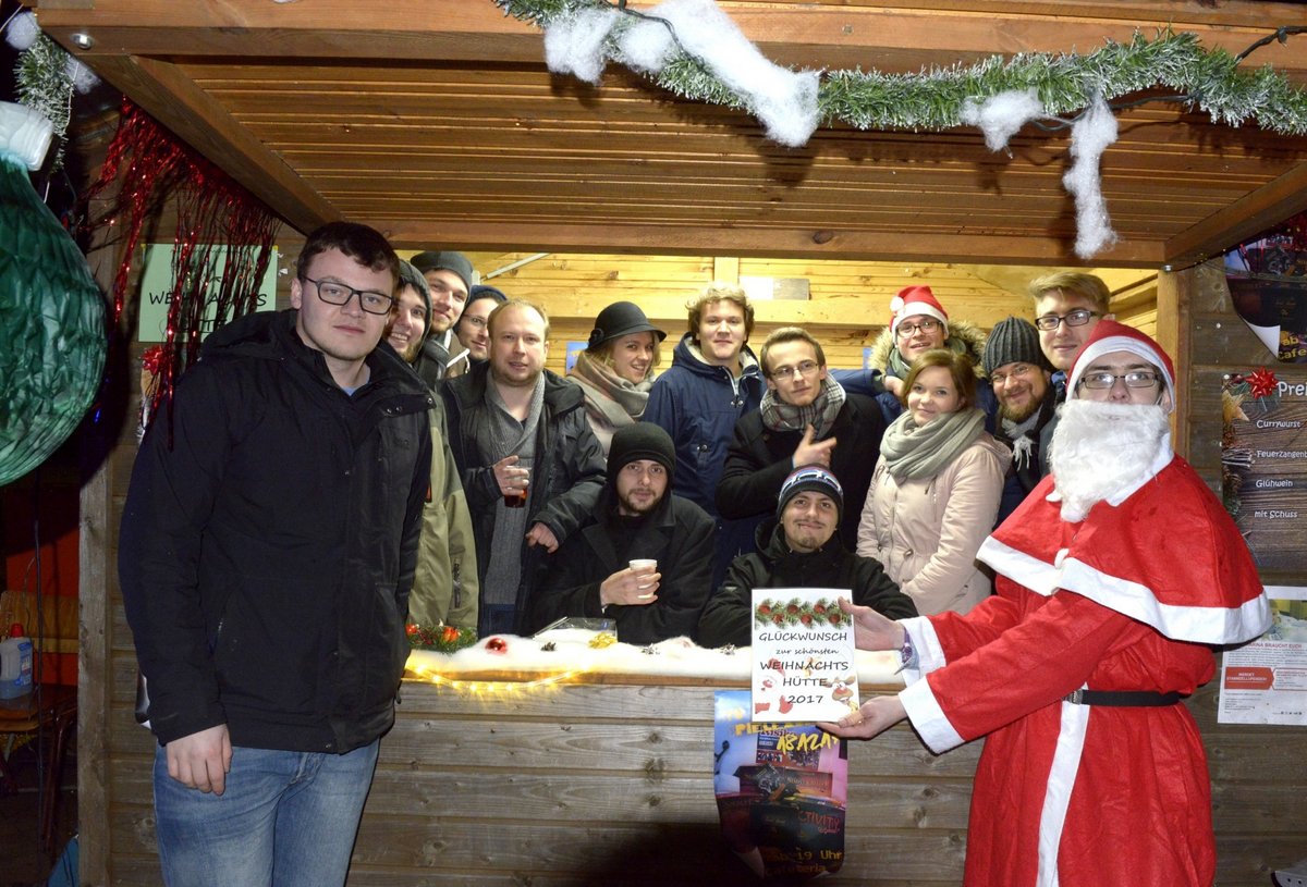 Das Siegerteam im Wettbewerb um die schönste Weihnachtshütte auf dem studentischen Weihnachtsmarkt 2017, in seiner Weihnachtshütte stehend, davor eine Person im Weihnachtsmannköstüm, welche die Urkunde für den 1. Platz präsentiert.
