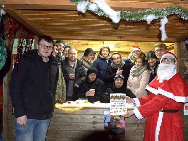 Das Siegerteam im Wettbewerb um die schönste Weihnachtshütte auf dem studentischen Weihnachtsmarkt 2017, in seiner Weihnachtshütte stehend, davor eine Person im Weihnachtsmannköstüm, welche die Urkunde für den 1. Platz präsentiert.