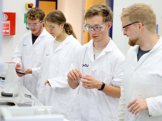 Eine Schülerin und drei Schüler stehen in weißen Kitteln in einem Chemielabor der BTU und experimentieren.