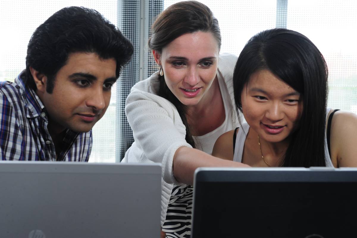 Drei Studierende sitzen vor zwei Laptops und erklären sich gegenseitig etwas