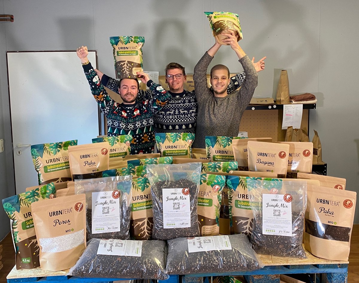 Drei Marketing-Absolventen der BTU Cottbus–Senftenberg freuen sich, hinter einem Tisch mit ihren Produkten stehend, über ihre Markteinführung. Foto: URBNTERA