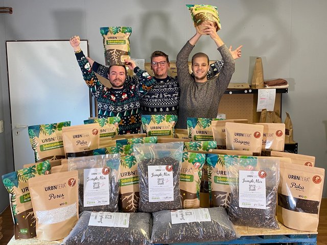 Drei Marketing-Absolventen der BTU Cottbus–Senftenberg freuen sich, hinter einem Tisch mit ihren Produkten stehend, über ihre Markteinführung. Foto: URBNTERA