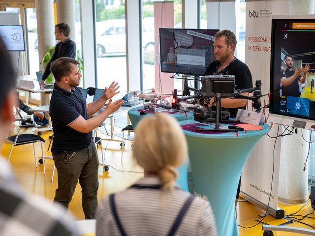 BTU-Mitarbeiter an einem Messestand geben Besuchern Erläterungen an einer Drohne zur zivielen Nutzung.  