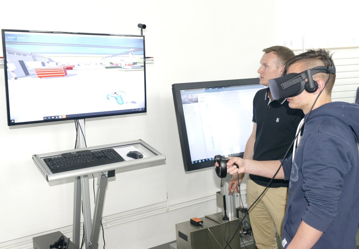 Ein Jugendlicher mit VR-Brille steht vor einem Monitor, der eine virtuelle Fabrik zeigt. Ein akademischer Mitarbeiter gibt neben ihm Erläuterungen.