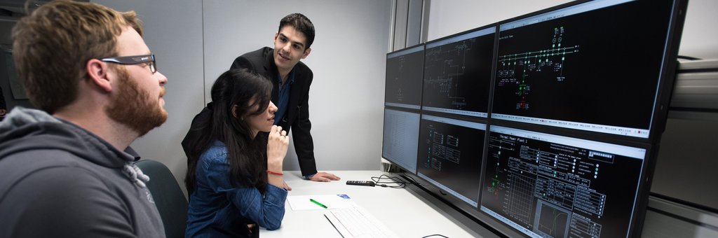 Umgang mit Hochspannungsnetzen am Lehrstuhl Energieverteilung und Hochspannungstechnik An einem Tisch, auf dem 6 Bildschirme in einem 2 mal 3 Raster angeordnet sind, sitzen 2 Studenten. Der Dozent steht am hinteren Tischende und spricht mit den Studenten.
