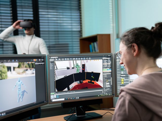 A student edits graphics on a PC, with a student wearing VR goggles in the background. Photo: BTU, Sebastian Rau
