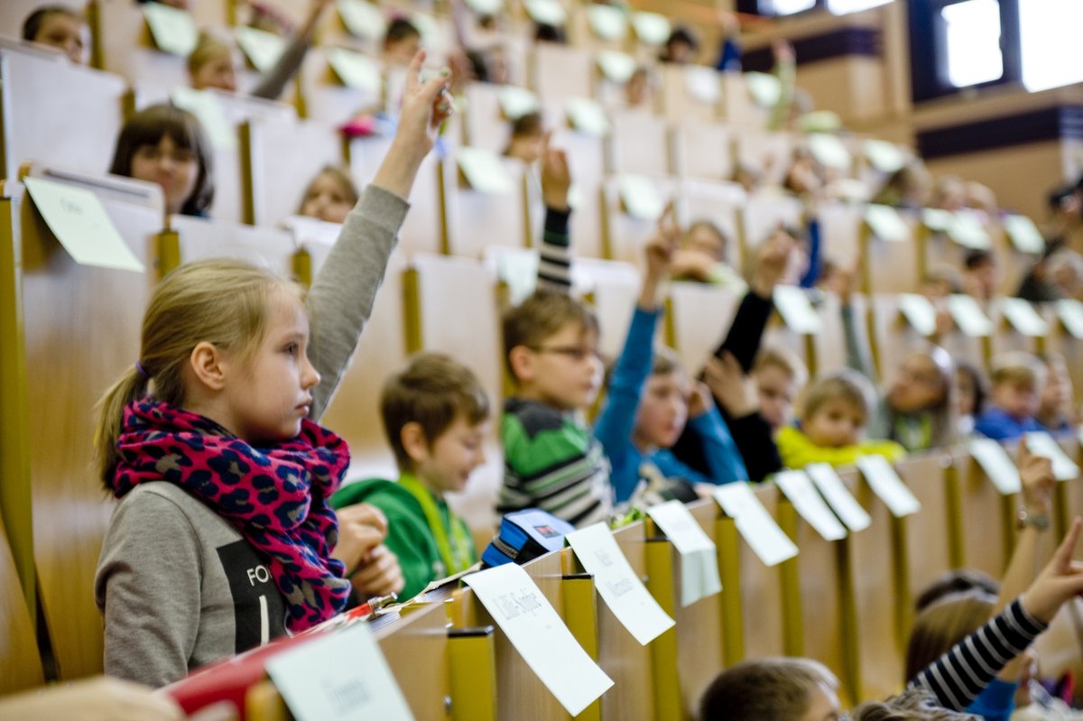 Mädchen und Jungen sitzen im Rahmen einer Kinderuni-Vorlesung in Hörsaalbänken und melden sich.