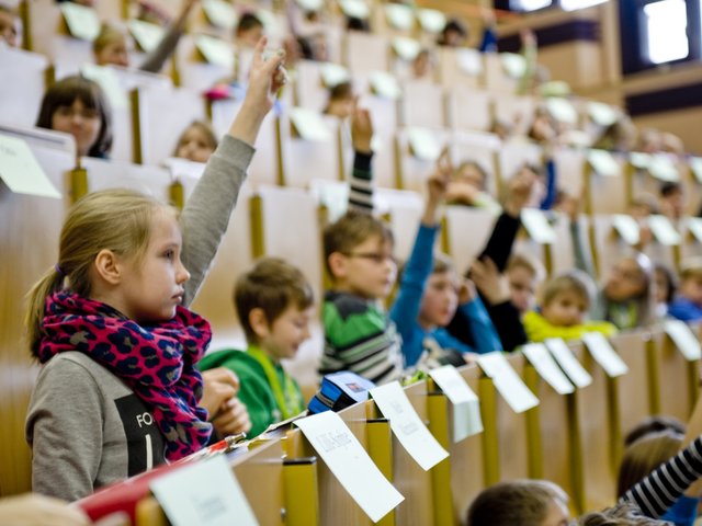 Mädchen und Jungen sitzen im Rahmen einer Kinderuni-Vorlesung in Hörsaalbänken und melden sich.