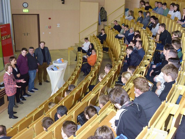 Studieninteressierte sitzen im Hörsaal und diskutiereren mit vor ihnen stehendne Professoren, Mitarbeitern und Unternehmensvertretern.