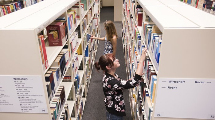 Das Foto zeigt eine Studientin, die in einem Bücherregal der Bibliothek ein Buch sucht.
