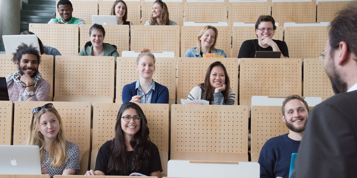 Studenten sitzen in einem Hörsaal. In der vorderen rechten Ecke ist der Dozent von hinten zu sehen.