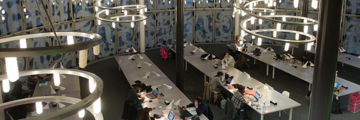 Draufblick auf einen abendlich dunklen Lesesaal im IKMZ. Im Vordergrund stehend lange Tische, an denen Studierende im Schein einzelner Lampen lernen. An der Decke hängen größe Spiralen mit Glühbirnen.Im Hitergrund sieht man durch die Glasfenster den Sonnenuntergang.