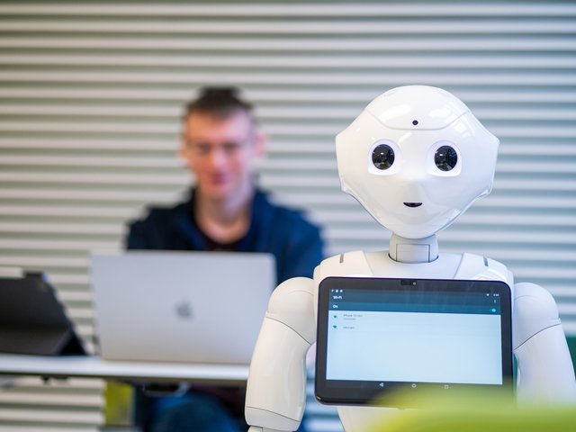Hinter eienm humanoiden Roboter arbeitet ein Student an einem Laptop.