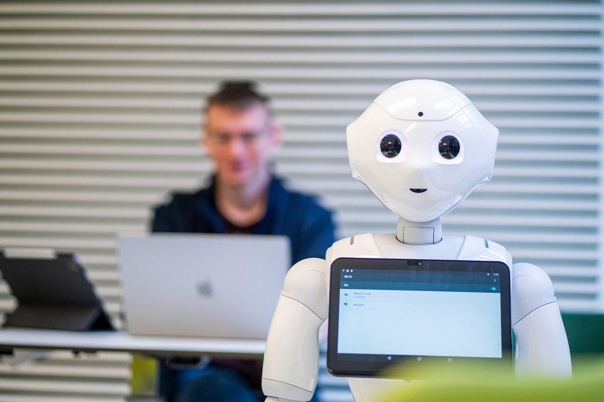 Hinter eienm humanoiden Roboter arbeitet ein Student an einem Laptop.