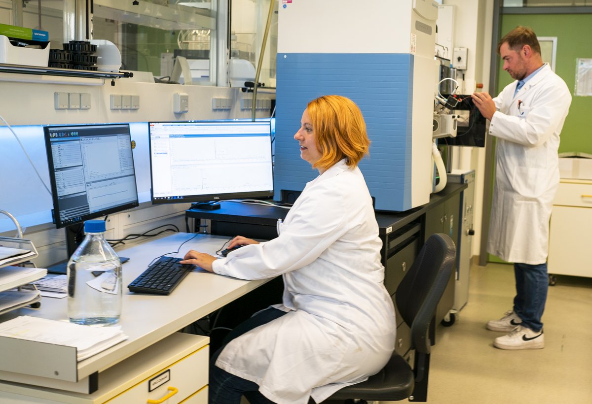 Eine Wissenschaftlerin wertet am PC Daten aus während im Hintergrund ein Wissenschaftler am Spektrometer arbeitet