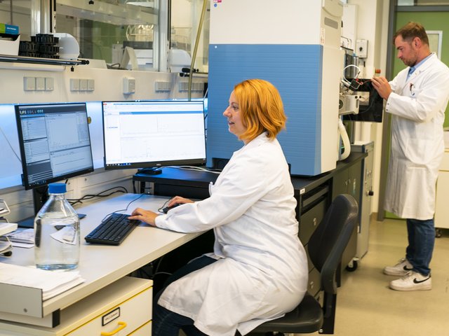 A scientist analyzes data on a PC while a scientist works on a spectrometer in the background.