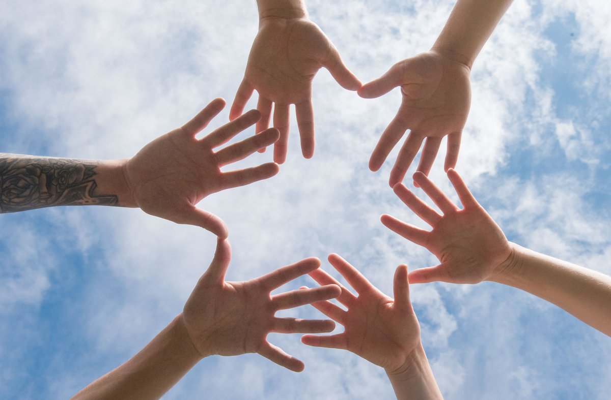 The photo shows 6 hands pictured in front of the sky, forming a circle.