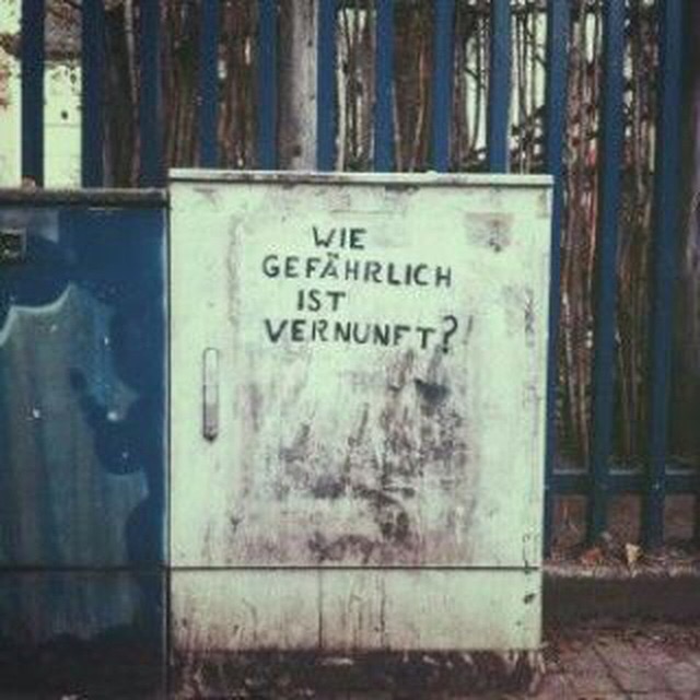 Die Grafik zeigt eine Fotografie. Im Fokus steht ein weißer Stromkasten mit einem Graffiti mit der in Großbuchstaben geschriebenen Frage „Wie gefährlich ist Vernunft?“. 