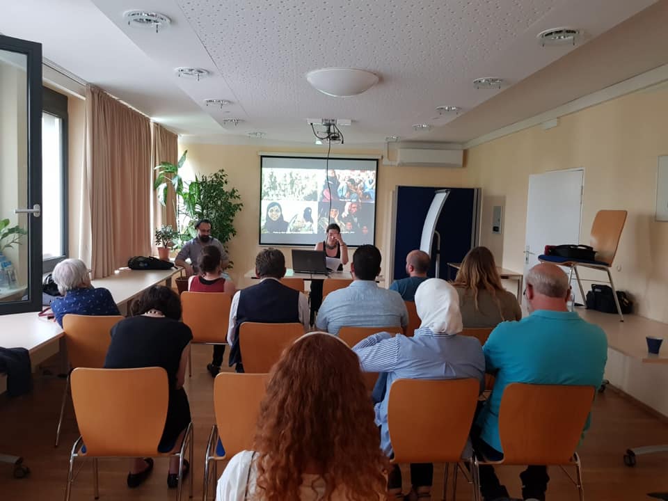 several people are sitting in an audience facing the female presenter and the whiteboard behind her with the presentation on it