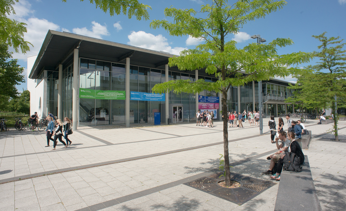 Studierende stehen vor dem Zentralen Hörsaalgebäude, andere sitzen auf Bänken vor dem Campus.