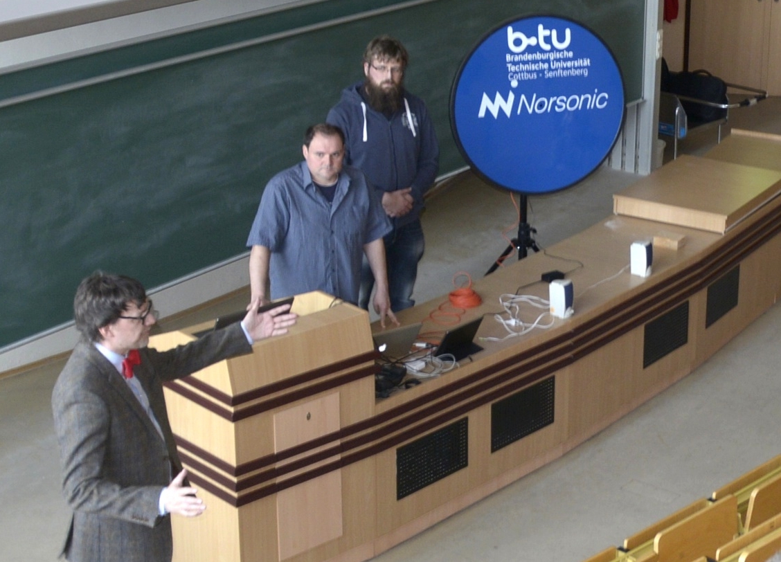Der wissenschaftliche Mitarbeiter Stephan Hernschier (Li.) und zwei weitere Personen geben, im Hörsaal sthend, Erläuterungen zur akkustischen Kamera.