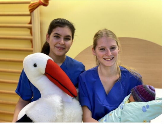 Das Foto zeigt zwei Studierende der Hebammenwissenschaft mit einem Plüschstorsch und einer Babypuppe im Arm. Beide lächeln.